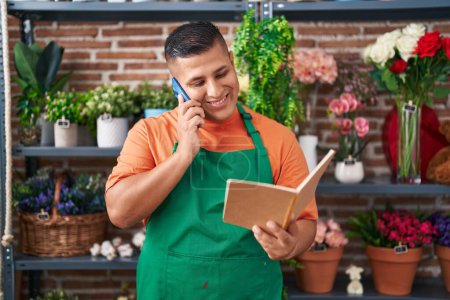 Photo for Young latin man florist talking on smartphone reading book at flower shop - Royalty Free Image