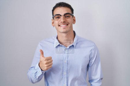 Foto de Hombre hispano guapo usando ropa de negocios y gafas haciendo un gesto feliz con la mano. aprobando la expresión mirando a la cámara mostrando éxito. - Imagen libre de derechos