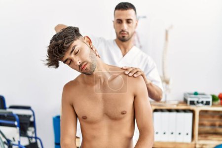 Foto de Dos hombres hispanos fisioterapeuta y paciente con sesión de rehabilitación estirando el cuello en la clínica - Imagen libre de derechos