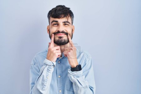 Téléchargez les photos : Jeune homme hispanique avec la barbe debout sur fond bleu souriant avec la bouche ouverte, doigts pointant et forçant sourire joyeux - en image libre de droit