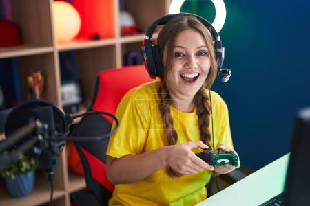 Photo for Young blonde woman streamer playing video game using joystick at gaming room - Royalty Free Image