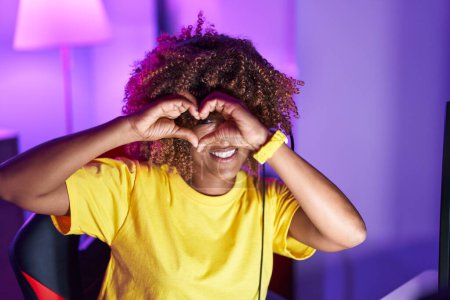 Téléchargez les photos : Femme afro-américaine streamer souriant confiant faire symbole de coeur avec les mains à la salle de jeux - en image libre de droit