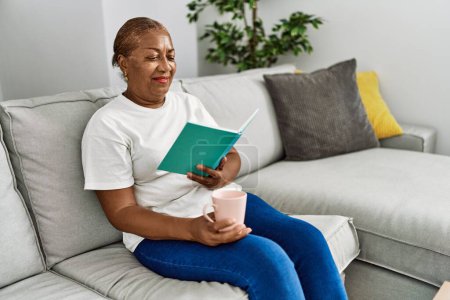 Foto de Mujer afroamericana mayor bebiendo libro de lectura de café en casa - Imagen libre de derechos