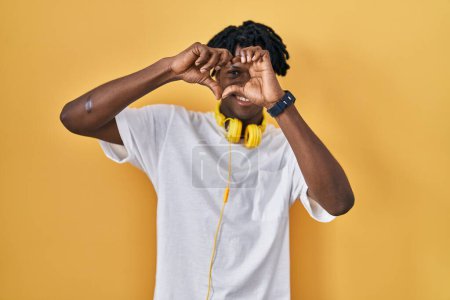Téléchargez les photos : Jeune homme africain avec dreadlocks debout sur fond jaune faisant forme de coeur avec la main et les doigts souriant en regardant à travers signe - en image libre de droit