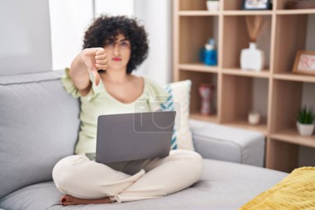 Photo for Young brunette woman with curly hair using laptop sitting on the sofa at home looking unhappy and angry showing rejection and negative with thumbs down gesture. bad expression. - Royalty Free Image
