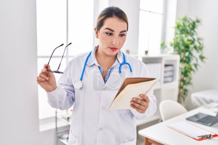 Photo for Young beautiful hispanic woman doctor reading notebook at clinic - Royalty Free Image