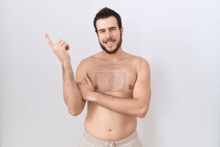 Téléchargez les photos : Jeune homme hispanique debout torse nu sur fond blanc avec un grand sourire sur le visage, pointant avec la main et le doigt vers le côté regardant la caméra. - en image libre de droit