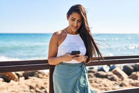 Téléchargez les photos : Jeune femme hispanique souriant confiant en utilisant un smartphone au bord de la mer - en image libre de droit