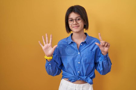 Foto de Chica joven de pie sobre el fondo amarillo que muestra y señala hacia arriba con los dedos número siete mientras sonríe confiado y feliz. - Imagen libre de derechos