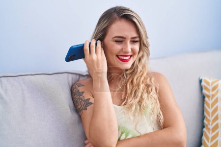 Téléchargez les photos : Jeune belle hispanique femme écoute un message vocal par smartphone assis sur le canapé à la maison - en image libre de droit