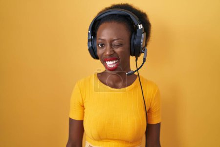 Foto de Mujer africana con el pelo rizado de pie sobre fondo amarillo con auriculares guiñando el ojo mirando a la cámara con expresión sexy, cara alegre y feliz. - Imagen libre de derechos