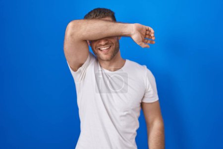 Téléchargez les photos : Jeune homme caucasien debout sur fond bleu couvrant les yeux avec le bras souriant gai et drôle. concept aveugle. - en image libre de droit