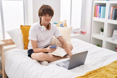 Téléchargez les photos : Jeune homme blond utilisant smartphone et ordinateur portable assis sur le lit dans la chambre - en image libre de droit