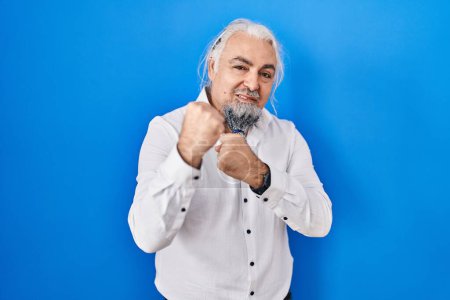 Photo for Middle age man with grey hair standing over blue background ready to fight with fist defense gesture, angry and upset face, afraid of problem - Royalty Free Image