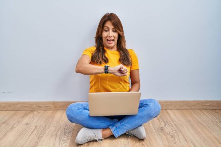 Foto de Mujer hispana usando laptop sentada en el suelo en casa mirando el tiempo del reloj preocupada, temerosa de llegar tarde - Imagen libre de derechos