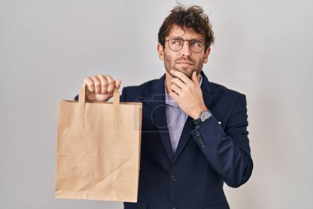 Photo for Hispanic business man holding delivery bag serious face thinking about question with hand on chin, thoughtful about confusing idea - Royalty Free Image