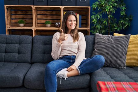 Photo for Beautiful woman sitting on the sofa at home smiling happy and positive, thumb up doing excellent and approval sign - Royalty Free Image