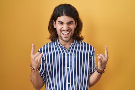Photo for Hispanic man with long hair standing over yellow background shouting with crazy expression doing rock symbol with hands up. music star. heavy concept. - Royalty Free Image