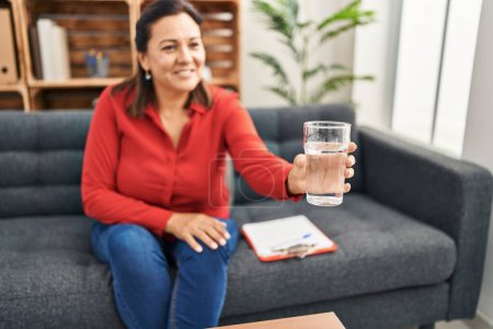 Photo for Middle age hispanic woman psychologist offering glass of water at psychology center - Royalty Free Image