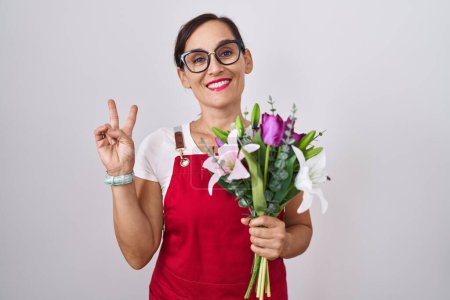Téléchargez les photos : Femme brune du moyen âge portant un tablier travaillant dans un magasin de fleuristes tenant un bouquet souriant avec un clin d'œil joyeux à la caméra faisant signe de victoire avec les doigts. numéro deux. - en image libre de droit