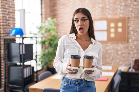 Téléchargez les photos : Jeune femme brune travaillant au bureau tenant à emporter café peur et choqué avec surprise et expression étonnante, peur et visage excité. - en image libre de droit