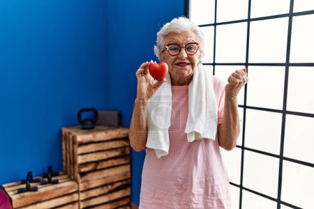 Photo for Senior woman with grey hair wearing sportswear holding red heart screaming proud, celebrating victory and success very excited with raised arm - Royalty Free Image