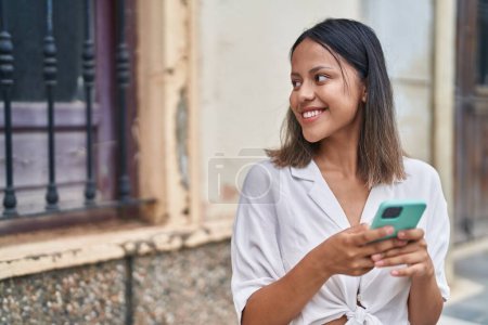 Foto de Joven mujer hispana sonriendo confiada usando smartphone en la calle - Imagen libre de derechos