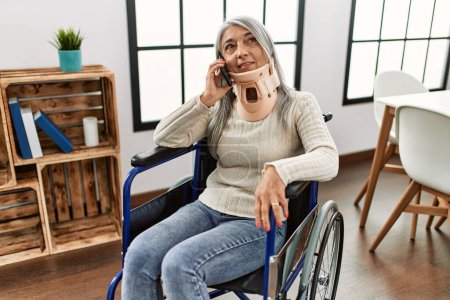 Téléchargez les photos : Femme aux cheveux gris d'âge moyen parlant sur le smartphone assis sur un fauteuil roulant à la maison - en image libre de droit