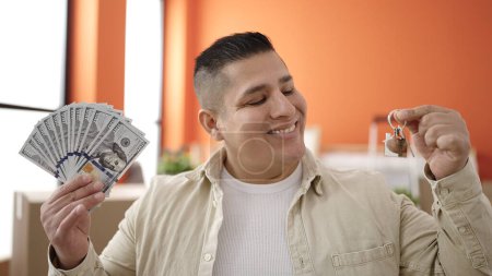 Téléchargez les photos : Jeune homme hispanique souriant confiant tenant de l'argent et des clés à la nouvelle maison - en image libre de droit