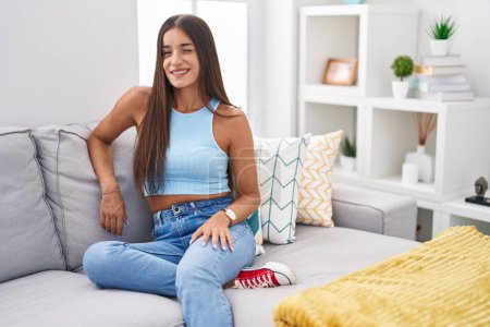 Téléchargez les photos : Jeune femme brune assise sur le canapé à la maison clignant des yeux en regardant la caméra avec une expression sexy, un visage joyeux et heureux. - en image libre de droit