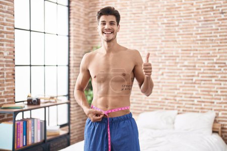 Photo for Young hispanic man using tape measure measuring waist smiling happy and positive, thumb up doing excellent and approval sign - Royalty Free Image