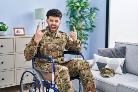 Photo for Arab man wearing camouflage army uniform sitting on wheelchair shouting with crazy expression doing rock symbol with hands up. music star. heavy concept. - Royalty Free Image