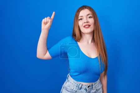 Téléchargez les photos : Femme rousse debout sur fond bleu montrant et pointant vers le haut avec le doigt numéro un tout en souriant confiant et heureux. - en image libre de droit