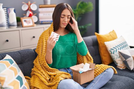 Photo for Young beautiful hispanic woman holding pills of delivery package sitting on sofa at home - Royalty Free Image