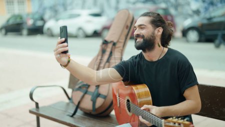 Photo for Young hispanic man musician holding classical guitar make selfie by smartphone at street - Royalty Free Image