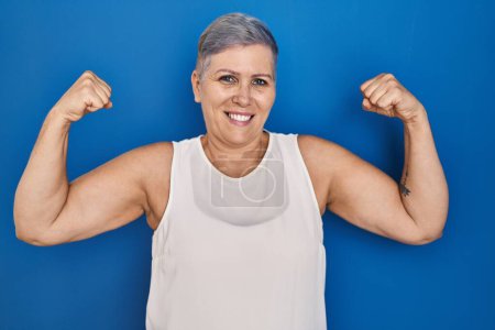 Photo for Middle age caucasian woman standing over blue background showing arms muscles smiling proud. fitness concept. - Royalty Free Image