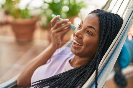 Photo for African american woman listening audio message by smartphone lying on hammock at home terrace - Royalty Free Image