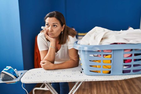 Photo for Middle age hispanic woman tired leaning on ironing board at laundry room - Royalty Free Image