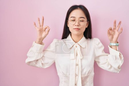 Téléchargez les photos : Jeune femme chinoise debout sur fond rose se détendre et sourire les yeux fermés en faisant geste de méditation avec les doigts. concept de yoga. - en image libre de droit
