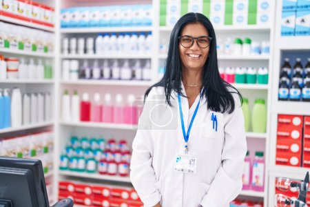 Foto de Farmacéutica hispana de mediana edad sonriendo confiada de pie en la farmacia - Imagen libre de derechos