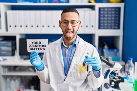 Foto de Un joven hispano que trabaja en el laboratorio científico sosteniendo sus asuntos de donación sosteniendo una muestra de sangre sacando la lengua feliz con expresión divertida. - Imagen libre de derechos