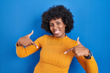 Foto de Mujer negra con el pelo rizado de pie sobre fondo azul mirando confiado con sonrisa en la cara, señalándose con los dedos orgullosos y felices. - Imagen libre de derechos
