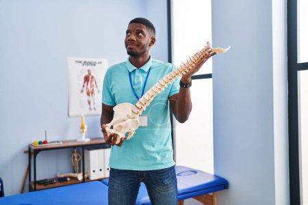 Photo for African american man holding anatomical model of spinal column smiling looking to the side and staring away thinking. - Royalty Free Image