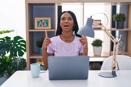 Photo for African american woman with braids using laptop at home amazed and surprised looking up and pointing with fingers and raised arms. - Royalty Free Image