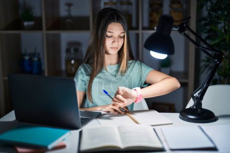 Téléchargez les photos : Adolescente faisant ses devoirs à la maison tard dans la nuit en vérifiant l'heure sur la montre-bracelet, détendue et confiante - en image libre de droit