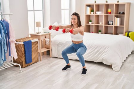 Foto de Joven hermosa mujer hispana entrenamiento pierna ejercicio usando mancuernas en el dormitorio - Imagen libre de derechos