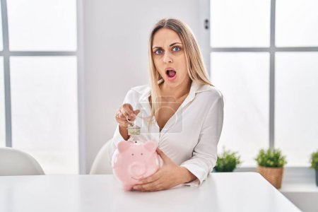 Photo for Young blonde woman holding piggy bank and house keys in shock face, looking skeptical and sarcastic, surprised with open mouth - Royalty Free Image