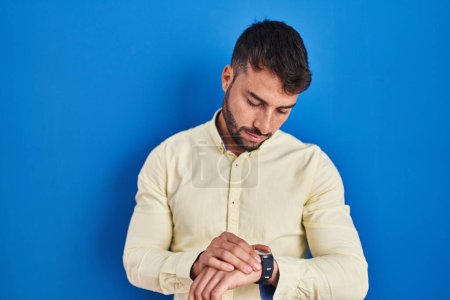 Téléchargez les photos : Bel homme hispanique debout sur fond bleu vérifier l'heure sur la montre-bracelet, détendu et confiant - en image libre de droit