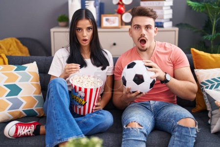 Photo for Young hispanic couple football hooligans holding ball and eating popcorn scared and amazed with open mouth for surprise, disbelief face - Royalty Free Image