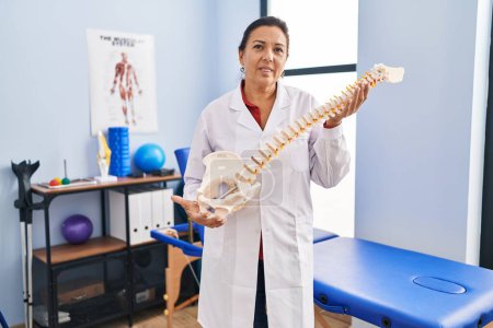 Photo for Middle age hispanic woman holding anatomical model of spinal column relaxed with serious expression on face. simple and natural looking at the camera. - Royalty Free Image